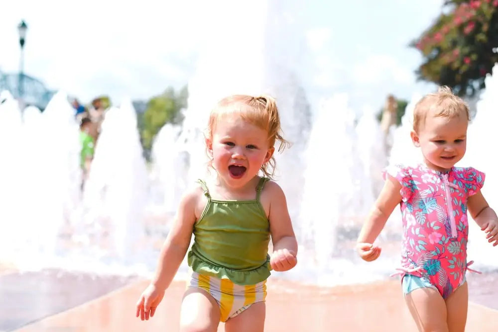 Baby girls in swimsuits next to a water fountain.